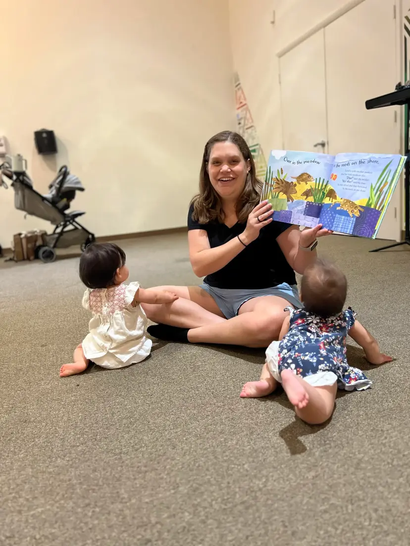 A grown-up shows a picture book to two babies sitting on the carpet